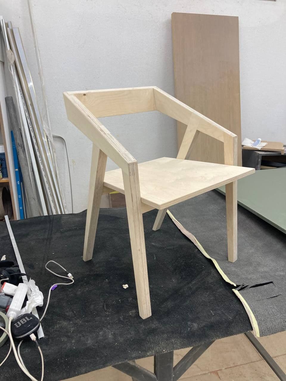 A modern wooden chair with angled legs sits on a dark surface in a workshop.